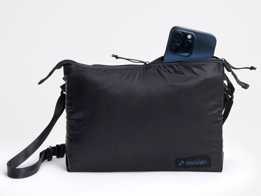 Pingora satchel in black with black straps and a small blue logo on the front, designed for carrying items while backpacking or traveling. Image shows size of bag compared to an iphone.