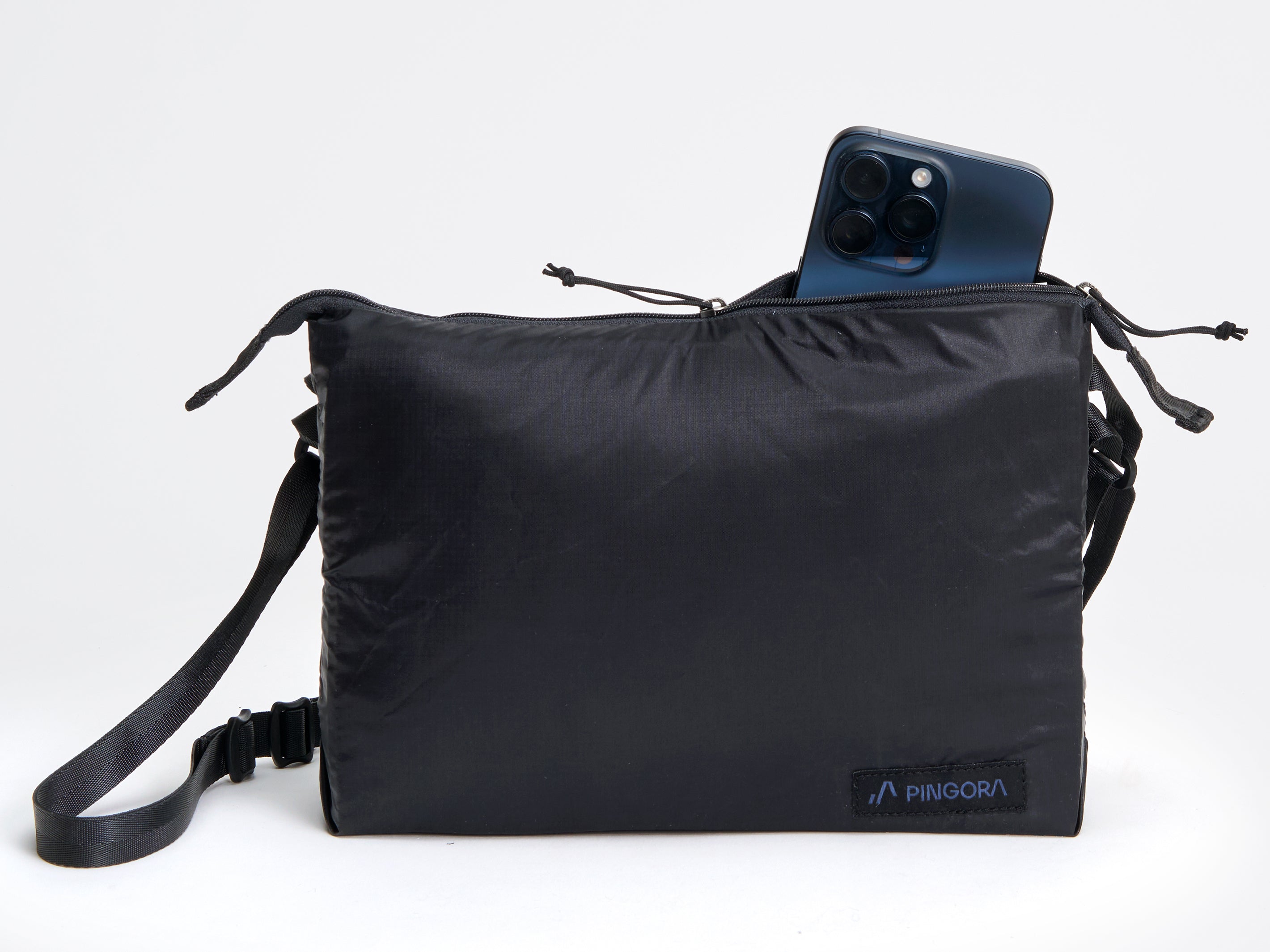 Pingora satchel in black with black straps and a small blue logo on the front, designed for carrying items while backpacking or traveling. Image shows size of bag compared to an iphone.